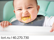 A dirty, joyful baby at the table on a feeding chair got his mouth dirty in vegetable puree. Introduction of complementary foods, the child likes to eat. Portrait close-up. Стоковое фото, фотограф Ольга Симонова / Фотобанк Лори