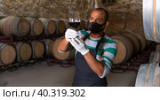 Owner in protective mask of winery standing with wine in wooden barrels cellar. Стоковое фото, фотограф Яков Филимонов / Фотобанк Лори