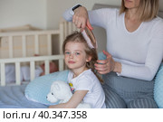 A happy young mother combs her adorable little daughter's hair while sitting in bed in the morning. The girl smiles. Family and care. Стоковое фото, фотограф Photojuli / Фотобанк Лори
