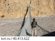 A man with a backpack stands on a suspension pedestrian bridge over a mountain gorge and looks at the mountains. Nepal. Himalayas. (2016 год). Стоковое фото, фотограф Михаил Пряхин / Фотобанк Лори