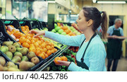 Positive middle-aged female market assistant in apron arranging tangerines on shelves in store. Стоковое видео, видеограф Яков Филимонов / Фотобанк Лори