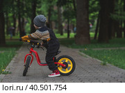 A cheerful little boy rides a bicycle outdoors. A happy child walks in the spring park. The kid is dressed in a fashionable sweater and denim overalls. Стоковое фото, фотограф Photojuli / Фотобанк Лори
