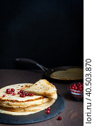 Stack of russian homemade traditional blinis or thin pancakes with red huckleberry served on round stone tray with pan ready for eating on dark brown wooden table against black background. Vertical. Стоковое фото, фотограф Елена Кабенкина / Фотобанк Лори