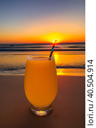 A glass of orange juice on the sea sand, small waves, romantic sunset. Стоковое AI-изображение, AI-художник Сергей Елизарьев / Фотобанк Лори