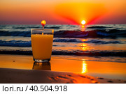 A glass of orange juice on the sea sand, small waves, romantic sunset. Стоковое AI-изображение, AI-художник Сергей Елизарьев / Фотобанк Лори