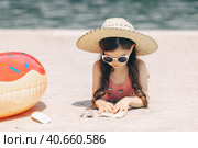 Happy cute smiling little girl in sunglasses lying on the sand on a beach in a swimsuit in a straw hat. Стоковое фото, фотограф Ирина Михайличенко / Фотобанк Лори