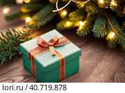 Green gift box with a gold ribbon on a wooden table next to a christmas tree with lights on it. Generative AI. Стоковое AI-изображение, AI-художник Mixail59 / Фотобанк Лори