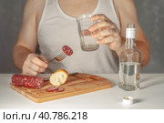 Алкоголик в майке пьет водку из граненого стакана и закусывает колбасой. A man, an alcoholic without a face in a white T-shirt, sits at a table and drinks vodka from a faceted glass, eating sausage and bread. Стоковое фото, фотограф Валерий Елистратов / Фотобанк Лори