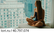 Young smiling woman drinking delicious coffee near window with big city skyscrapers outside. Relaxed girl enjoys morning cappuccino on weekend in the metropolis. Slow motion close-up footage. Стоковое фото, фотограф Ольга Сухарева / Фотобанк Лори