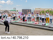 Пенсионеры, участники спортивного праздника программы "Московское долголетие", в фирменных московских футболках и шапочках, стоя на разноцветных гимнастических ковриках, выполняют гимнастику под руководством опытного тренера в парке Музеон на фоне московских зданий и голубого неба с красивыми белыми облаками (2023 год). Редакционное фото, фотограф Сергей Никифоров-Гордеев / Фотобанк Лори