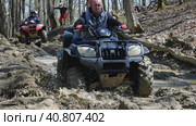 A couple on a black and red ATV ride through mud in the spring forest. Extreme type of outdoor activities. Touring trips on a quad bike (2022 год). Редакционное фото, фотограф Воловодов Владимир / Фотобанк Лори