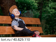 Cute, cheerful, little boy smiles and listens to music in big blue headphones in nature in the park. Music for children's development. Стоковое фото, фотограф Photojuli / Фотобанк Лори