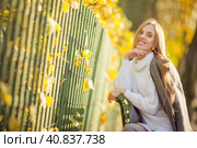 Beautiful happy girl with light brown hair is sitting on a bench near the fence in the autumn park. Attractive young woman in a gray coat and white sweater is smiling. Fall season. Autumn mood. Стоковое фото, фотограф Андрей Степанов / Фотобанк Лори
