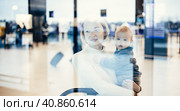 Thoughtful young mother looking trough window holding his infant baby boy child while waiting to board an airplane at airport terminal departure gates. Travel with baby concept. Стоковое фото, фотограф Matej Kastelic / Фотобанк Лори