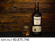 A bottle of Dewar's 12 years old Scotch whisky and a glass with ice on a dark wooden background. One of the most titled whiskies of its kind in the world. (2021 год). Редакционное фото, фотограф Воловодов Владимир / Фотобанк Лори