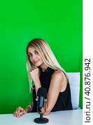 Lovely woman broadcaster with microphone sitting at desk at green background, looking at camera. Portrait of female broadcaster posing in television studio. Tv production concept. Copy ad text space. Стоковое фото, фотограф Aleksandr Golubev / Фотобанк Лори