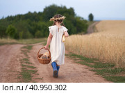 A girl with a bread basket in one hand and a long bread in the other hand is walking along a field road through a wheat field. She is wearing a linen dress and a wreath of spikelets on her head. Стоковое фото, фотограф Роман Дзюбало / Фотобанк Лори
