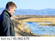Young man in coat looking down with upset face near swamp and rail. Depressive concept loss of money, loneliness, unemployment. Copy space. Стоковое фото, фотограф Татьяна Терехина / Фотобанк Лори