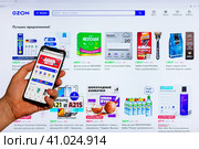 Adygea, Russia - August 09, 2023: hand man with smartphone making purchases in the Ozon online store through mobile application against background of Ozon marketplace web page open on computer monitor. Редакционное фото, фотограф Андрей С / Фотобанк Лори
