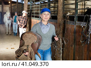 Elderly female rancher walking in stable with saddle in hands. Стоковое фото, фотограф Яков Филимонов / Фотобанк Лори