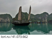 An old sailing boat stands on the smooth water of Halong Bay. The beige sails are worn out, with holes. Picturesque islands with steep slopes against a foggy sky. Fishing water houses in the distance. Стоковое фото, фотограф Вера Ивановна Кубарева / Фотобанк Лори