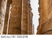 Colonnade in the hypostyle hall of the Karnak Temple in Luxor. A narrow passage between rows of columns. Hieroglyphs and carvings are visible. Ahead, against the sky - an obelisk. Egypt (2021 год). Стоковое фото, фотограф Вера Ивановна Кубарева / Фотобанк Лори