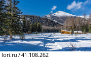 The winter road is trampled in the valley. In the distance, the wooden houses of the tourist base are visible. Picturesque wooded mountains against a blue sky. Coniferous trees in the snow. Altai. Стоковое фото, фотограф Вера Ивановна Кубарева / Фотобанк Лори