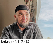 Portrait of senior asian man with beard wearing skullcap sitting by window. Стоковое фото, фотограф Ирина Кононова / Фотобанк Лори
