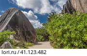 A pedestrian path passes between huge granite boulders. Lush tropical vegetation on the roadsides. Blue sky with clouds. Seychelles. La Digue Island. Стоковое фото, фотограф Вера Ивановна Кубарева / Фотобанк Лори