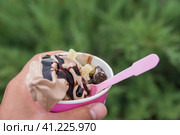 Closeup photo of paper cup with ice cream in man hand over green background in the park. Coffee ice cream with candied fruits and chocolate cookies. Side view with selective focus. Фотография крупным планом бумажного стаканчика с мороженым в руке мужчины на зеленом фоне в парке. Кофейное мороженое с цукатами и шоколадным печеньем. Вид сбоку с выборочной фокусировкой. Стоковое фото, фотограф Дмитрий Березнев / Фотобанк Лори