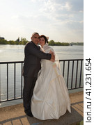 The bride and groom are hugging on the embankment of the river. A woman is dressed in a white dress, a man in a suit. Sunny weather over the river. Стоковое фото, фотограф Юлия Петровская / Фотобанк Лори