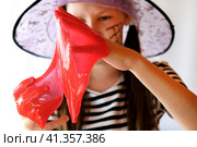 Cute girl playing with a red bloody glitter slime. She is having fun and making faces. Adorable girl holding and stretching a red goo slime. Halloween celebration, masquerade, trick or treat concept. Стоковое фото, фотограф Марина Демидюк / Фотобанк Лори