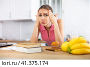 Young woman with headache standing near table with books at home. Стоковое фото, фотограф Татьяна Яцевич / Фотобанк Лори