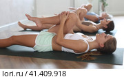 Relaxed sporty teen girl doing yoga with group in studio, lying on mat with knee pressed to chest with hands, performing hamstring and low back stretching Ardha Pawanmuktasana. Стоковое видео, видеограф Яков Филимонов / Фотобанк Лори