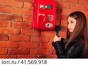 Pensive child girl in leather jacket holding handset old red payphone at brick wall, thought looking away. Cute kid lady model posing near telephone. Support communicate concept. Copy ad text space. Стоковое фото, фотограф Aleksandr Golubev / Фотобанк Лори