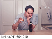 A man looks thoughtfully at a bottle of alcohol sitting in the kitchen with a glass in his hand. Problems of alcoholism in loneliness, sadness and melancholy. Стоковое фото, фотограф Андрей Журавлев / Фотобанк Лори
