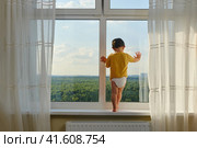 The child is precariously perched on the window sill, looking out the window and risking a fall. Kid aged two years. Стоковое фото, фотограф Андрей Журавлев / Фотобанк Лори