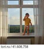Baby is standing on the window sill, gazing out at the sky and risking a life-changing accident. Kid aged two years. Стоковое фото, фотограф Андрей Журавлев / Фотобанк Лори