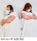 A man and a woman in medical masks are angry at each other. The husband and wife lie turned away in bed on isolation from the coronavirus quarantine. Стоковое фото, фотограф Андрей Журавлев / Фотобанк Лори