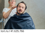 A woman cuts a man's hair at home and injures his ear, copy space. Concept of closed hairdressers and barbershops in isolation from the epidemic of the coronavirus. Стоковое фото, фотограф Андрей Журавлев / Фотобанк Лори