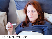 Woman with a coronavirus infection in a medical mask looks at the thermometer in surprise. Стоковое фото, фотограф Андрей Журавлев / Фотобанк Лори