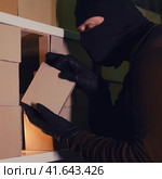 A thief in a black hood steals a parcel in a warehouse in the dark. Concept of problems with theft of goods and postal parcels. Стоковое фото, фотограф Андрей Журавлев / Фотобанк Лори
