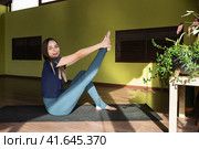 Молодая улыбающаяся спортивная девушка шатенка выполняет динамическую растяжку мышц, сгибающих ногу в колене, тренируется в спортивной одежде сидя на коврике в современной фитнес-студии. Стоковое фото, фотограф Сергей Чайко / Фотобанк Лори