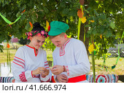 Клязьминский городок (Стародуб), Владимирская область, Россия. Август 2023 года. Юноша и девушка в ярких народных костюмах с улыбкой читают записку под зеленью дуба. Редакционное фото, фотограф Сергей Никифоров-Гордеев / Фотобанк Лори