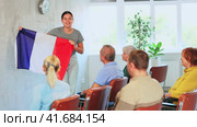 Positive friendly young female tutor giving lecture to group of older adults during educational class, holding national flag of France and talking about history of country. Стоковое видео, видеограф Яков Филимонов / Фотобанк Лори