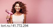 A young beautiful girl stands with a hair dryer on a pink monochrome background. Copy space. Стоковое AI-изображение, AI-художник Ярославский Максим / Фотобанк Лори