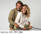 A young couple stands on a gray background with a supermarket cart and smiles. Стоковое AI-изображение, AI-художник Ярославский Максим / Фотобанк Лори
