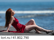 Woman in red velvet long dress with raised hem and bare legs lies on her back on black sand on beach. Стоковое фото, фотограф А. А. Пирагис / Фотобанк Лори