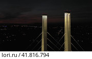 A cable-stayed highway bridge with tall concrete pylons illuminated by architectural lighting at night. Vertical climb up along the pylons against the background of night city lights with camera rotation. (2023 год). Редакционное видео, видеограф Даниил Хабаров / Фотобанк Лори