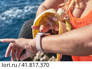 Latin woman, middle-aged, resting, regaining strength, eating, drinking water, after a gym session, burning calories, keeping fit, outdoors by the sea, wearing headphones and smart watch. Стоковое фото, фотограф carlos l vives / Фотобанк Лори